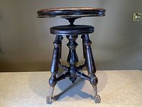 Front view of the antique adjustable piano stool showing the round wooden seat, turned legs, and metal ball and claw feet with casters.