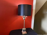 Front view of the full lamp showing the black silk drum shade, metal stem with palm tree decoration, and lucite base.