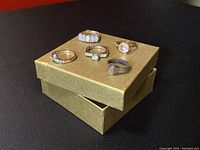 Five assorted rings displayed on a gold-colored box top showing various designs and mixed materials.