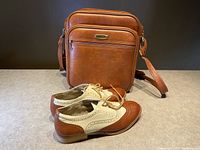 Eaton brown leather shoulder bag displayed standing upright with size 10 two-tone brown and cream bowling shoes in front, on a gray surface against black background.