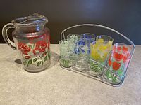 Full view of glass pitcher with red rose design next to 8 floral printed glasses in silver metal caddy.