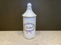 Front view of white ceramic apothecary jar with 'Hashish' label and colorful floral wreath decoration