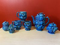 Photo showing full set of ceramic tea set including teapot, three mugs, sugar jar, jam jar and shaker, all with detailed relief fruit decoration in blue and green glaze.