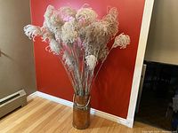 Photo showing full view of vintage copper milk can vase filled with pampas grass against a reddish wall.