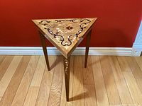 Front view of triangular wooden accent table showing detailed floral and scroll wood inlay on top surface and sides.