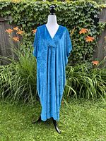 Blue vintage night gown displayed on a mannequin outside against a backdrop of green foliage and orange flowers, showing the full front view with loose fit and floral embossed velvet fabric.