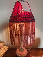 Front view of the table lamp illuminated showing red beaded shade with hanging bead strands and ornate white base.