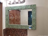 Rectangular wall mirror with a frame of green distressed tiles embracing a geometric pattern, photographed hanging on a bathroom wall above a tiled backsplash.