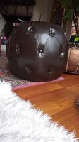 Side view of dark brown faux leather tufted ottoman on wooden floor with area rug and plant in background.