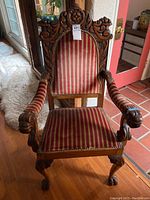 Front view showing red and gold striped velvet upholstery, hand-carved wood details on the high backrest, armrests ending in carved lion heads, and claw feet on legs.