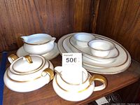 Photo of porcelain serving set including sugar bowl, creamer, gravy boat with plate, and oval serving dishes arranged on wood shelf.