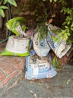 Five partial bags of mulch and potting soil, displayed outdoors on stone and brick surfaces surrounded by plants.