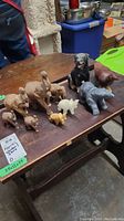 Photo showing multiple animal figurines on wooden table including wooden elephants, a black ceramic dog, gray stone hippopotamus, white soapstone polar bear.