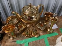 Overview photo of all eleven reflective gold-glazed china pieces arranged on a wooden surface, showing the teapot, condiment containers, candle holders and bud vase.