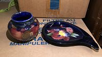 Photo of the Moorcroft fruit themed bud vase and floral ash tray showing the overall shapes and colors of both items on top of cardboard boxes.