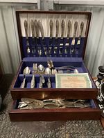Full view of open two-tier wooden flatware case showing knives held upright in slots on the inside lid, assorted spoons, forks, and serving utensils in compartments below