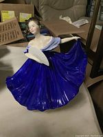 Front and slight side angle showing the figurine holding a white fan, cobalt blue dress with off-the-shoulder sleeves, and curled hair with a yellow ribbon.