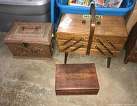 Three wooden sewing boxes displayed on floor with blue storage bins behind. Boxes include an accordion style, a smaller ornately carved box, and a medium-sized box.