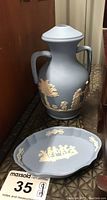 Photo showing blue Wedgwood Jasperware lamp base with two handles and classical white relief figures, standing behind blue and white Jasperware trinket dish with white relief children motif.