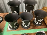 Five black jasperware cups with white cherub and angel reliefs arranged on wooden table.