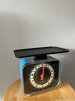 Front view of blue metal kitchen scale with black weighing tray on top, showing dial with white markings and red needle pointer.
