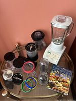 Photo showing Magic Bullet blender base with blade, black tamper tool, multiple clear blending cups and lids, colored rubber rings, larger blender pitcher, white second blender, and recipe book on a round glass table.