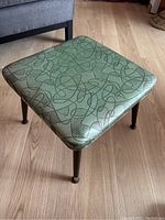 Square vinyl-upholstered ottoman with green abstract atomic lines pattern on textured surface on a wood floor.