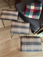 Three neatly stacked Mid-Century Modern style stools with metal frames and geometric fabric cushions, shown on a wood floor near a dark upholstered chair with the folded multicolored checkered fleece blanket on it.