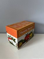 Side perspective of the metal recipe box showing orange lid and vegetable illustrations on the side.
