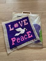 Full needlepoint piece on canvas with purple yarn, pink letters, white dove with flower, and yarn in plastic bag.