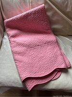 Pink quilted blanket folded on a beige velvet couch, showing texture and quilted pattern details.