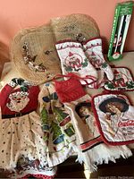 Photo shows various Christmas-themed oven mitts, dish towels and vintage Coca-Cola kitchen linens arranged on a table, some wear visible.