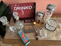 Overview of vintage barware items including cut glass decanters, DRINKO game, metal cocktail shaker, ice bucket, muddler, cocktail fork set, bottle opener and boxed siphon jigger.