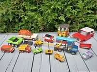 Full view of collection of 14 vintage toy vehicles including construction vehicles, trucks, small cars, and a toy drum labeled 'Tiny Town'. Colors vary including green, red, yellow, blue, orange, and purple. Items arranged on wooden surface in front of green hedge.