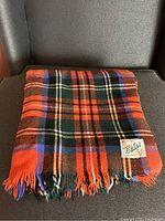 Folded plaid wool blanket with strong orange, brown, green, purple and cream colors arranged in a traditional tartan pattern placed on a dark surface.