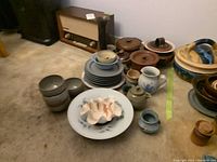 Wide view of assorted pottery pieces including bowls, plates, vases, pots, and stacked dishes on floor near vintage radio.