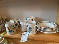 Five ceramic mugs and three ceramic bowls on a wooden shelf with botanical flower designs, floral prints, and green leaf bordered rims.