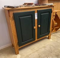 Front view of wood cabinet showing natural finish frame and two dark green paneled doors with wood knobs. Shows wear on surfaces.