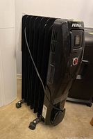 Black Noma oil filled heater, side perspective showing fins, caster wheels, and attached power cord.