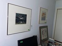 Wide view of all three framed original art pieces hanging on the wall above a desk with computer and papers, showing their layout and framing.