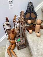 Photo shows wooden artist model sitting with one arm raised, in front of miniature twig chair sculptures and a rustic paper sculpture figure. Large pulley and other wooden figures visible in background.