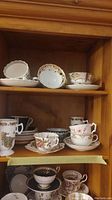 Wide view showing multiple teacups and saucers arranged on wooden shelves in a cabinet, variety of patterns and styles visible