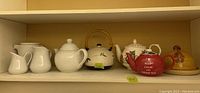 Shelf view showing six ceramic items including four teapots, two creamers, and cheese dome