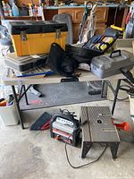 Full view of table with multiple toolboxes, a battery charger on the ground, a broom, and a small portable table saw