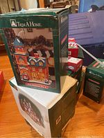 Photo showing three boxed Christmas village houses, including 'Fire Station No. 6' by Trim A Home, stacked on and around a table.