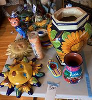 Wide view of the pottery lot on table, showing large sunflower vase, sunflower face, sun decor, colorful vase, candle holder, painted cat figurine, and small bowl