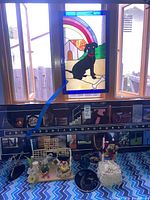 Stained glass panel featuring black dog with rainbow background hanging in window above patterned surface with assorted figurines and decor.