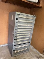 Front view showing all 12 drawers with white handles and labels on a gray metal cabinet placed against a peach wall.