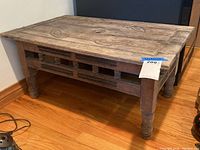 Front view of a rectangular carved wood table on hardwood floor, showing top surface and carved spindle accents below.