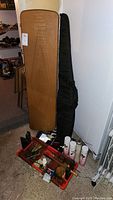 Full view of brown hardsided guitar case with black soft gig bag behind it and items on floor in front, includes toolbox and aerosol cans.